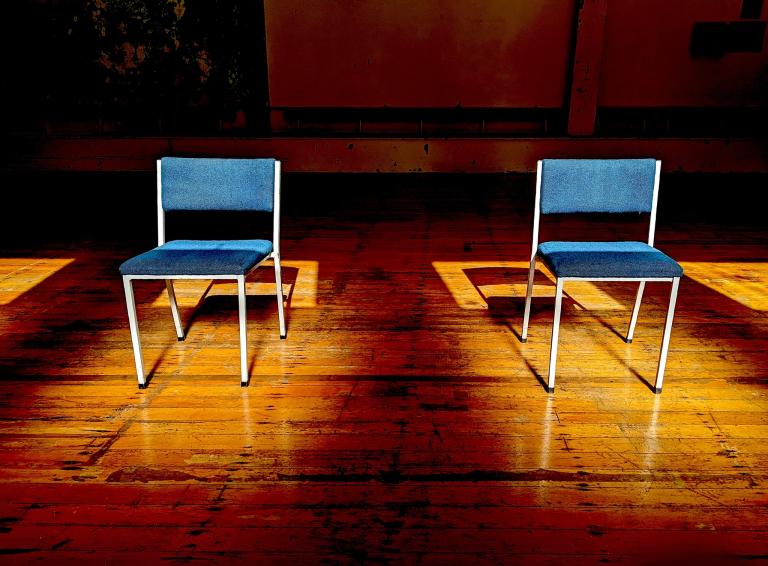 two empty chairs on in a sunbeam. The floor is wood and is possibly in a school hall.