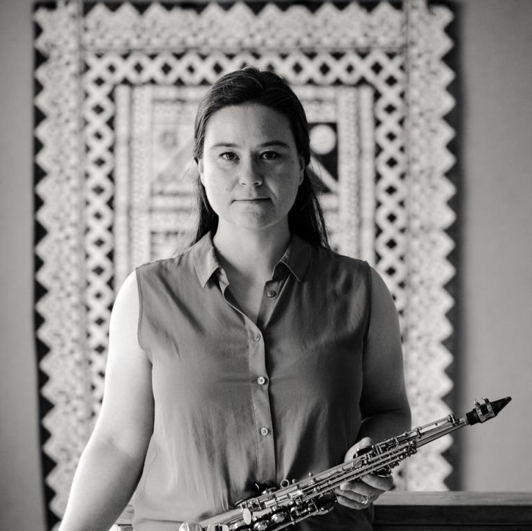 A black and white photo featuring Jasmine Lovell-Smith standing in the centre holding a soprano saxophone with a patterned tapestry hanging in the background