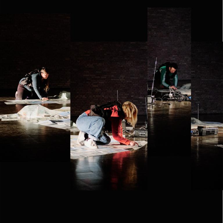 LTK4 performing on the floor of a dark stage with 1980/1990s radios, sheets of paper draped over the floor. The performers appear in work vests strapped around their torsos. 