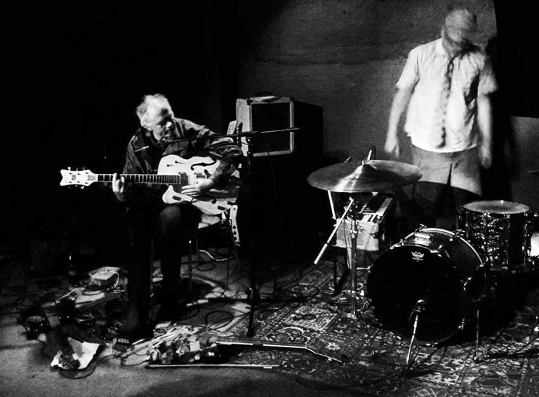 Black and white photo of Greg Malcolm hunched over his guitar and pedals and a blurry Stefan Neville looming over his drumset