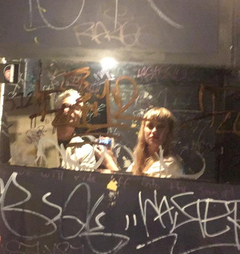 two people stand in front of a dirty graffiti-covered venue bathroom mirror taking a selfie