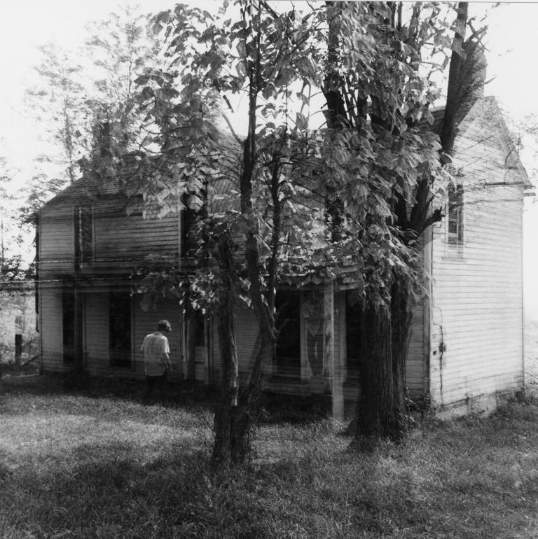 a black and white photo of an old house. The exposure of the photo was clearly long, the house is doubled and ghostly figures stand in various positions in the photo. Photo by Eugene Meatyard