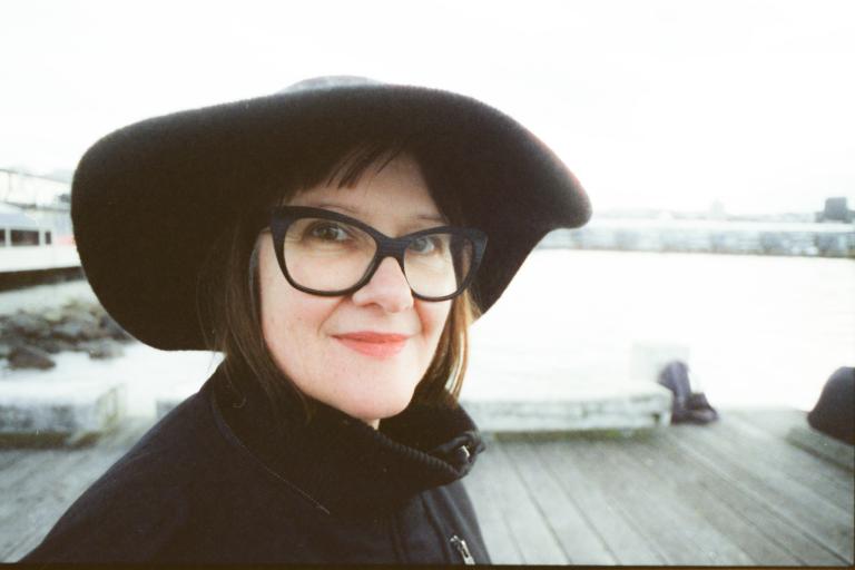 Sonya Waters smiles at the camera in a black hat and black outfit on a pier on the sea