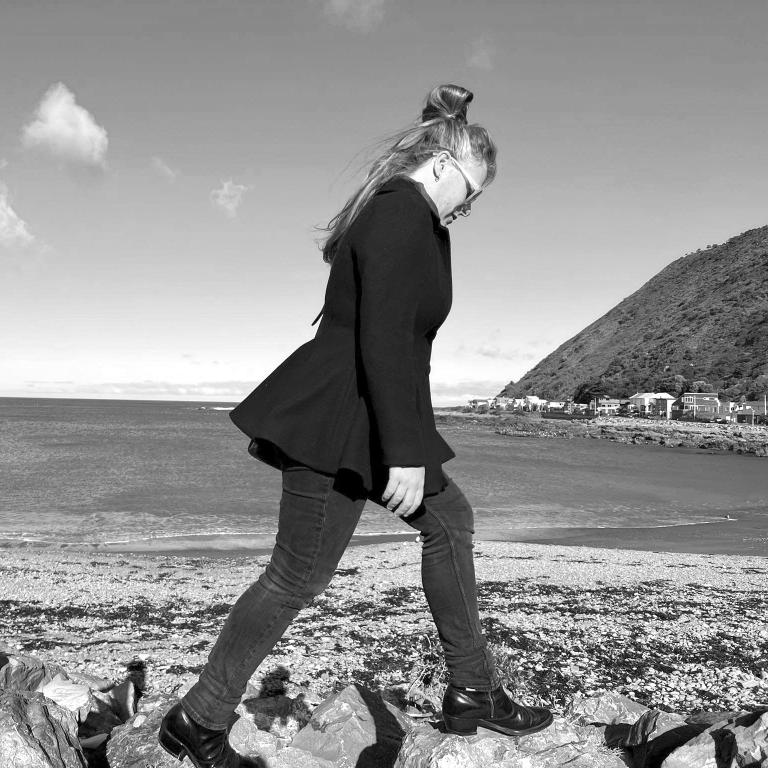 A black and white photo of Rosie Whinray. She walks carefully, one foot at a time along the rocks of a stoney beach. There is a hill cascading down into the sea on the right, off into the distance. The photo is likely taken along the south coast in Wellington, where Rosie collected much of the material for her Pyramid Club exhibition. It is a bright day with few, small clouds. 