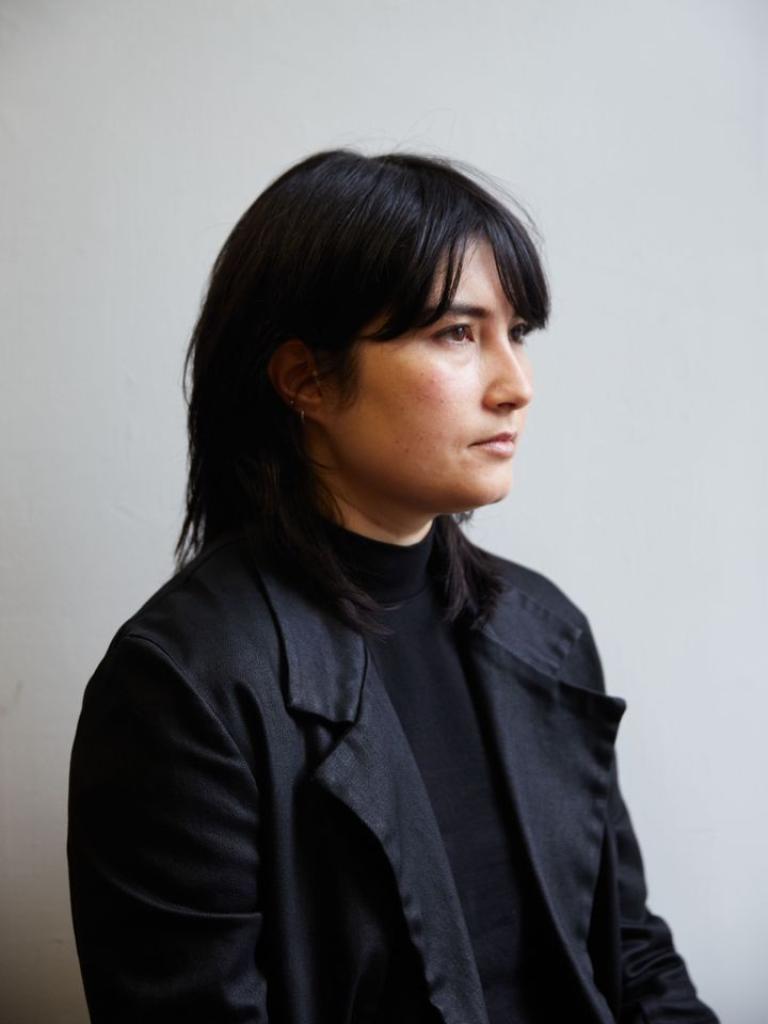 Emi Pogoni sits in front of a grey wall. They are wearing all black and the light is softly catching their face.