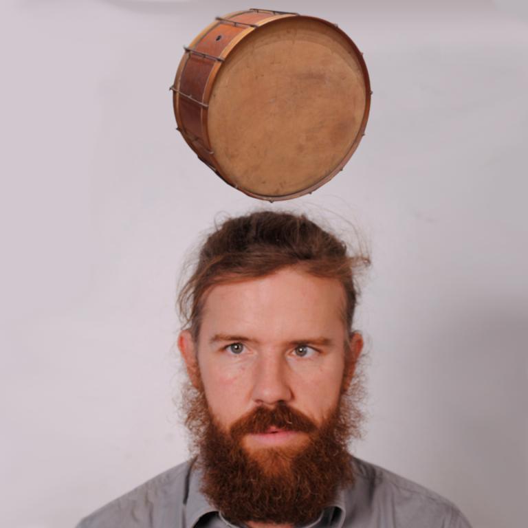 A front-on portrait of Chris O'Connor from the shoulders up. Above Chris' head a drum levitates.
