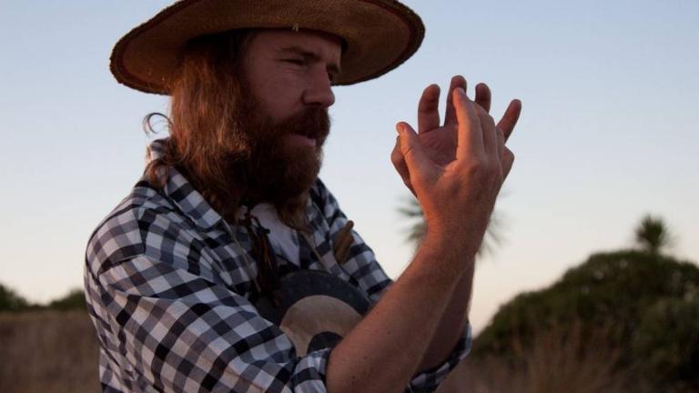 Chris O'Connor stands in front of a sunset explaining something with his hands. He's wearing a large brim hat