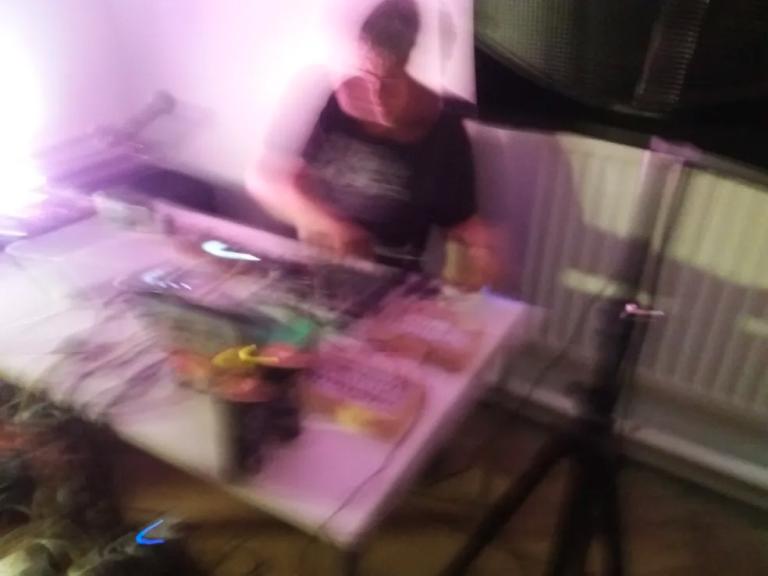 a blury digital camera photo from above an s glass set. S glass has objects arranged across the table including blury electronics. a purple light washes out the left side of the photo. S Glass looks out of breath in an intense moment of performance. 