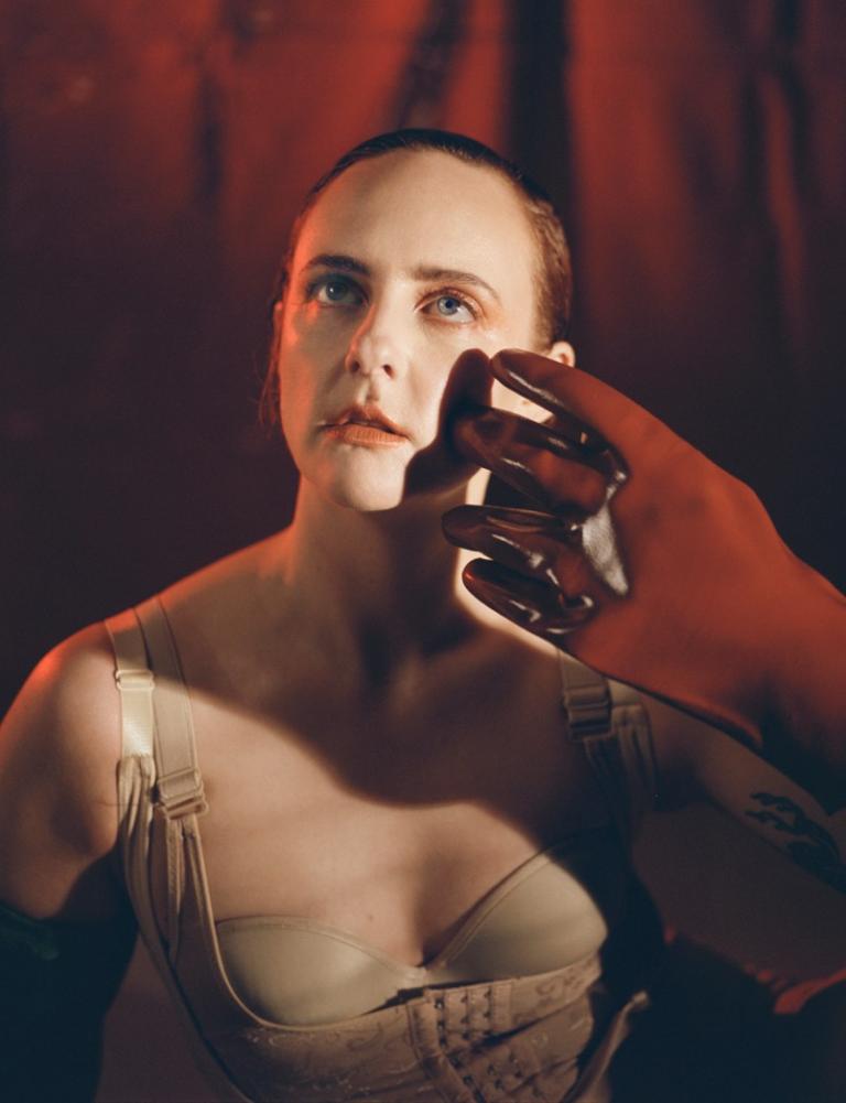 A front-on portrait style photo of i.e. crazy in front of a red curtain background. A red rubber glove reaches out and strokes the side of their face. They are wearing a cream coloured corset and are looking upwards off into the distance, the photo has a dreamy surreal vibe about it. 