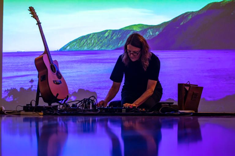 Jacqui O'Reilly sitting down performing in front of a projection of the ocean and a mountain