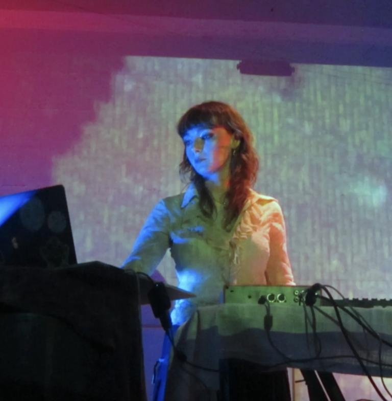 Fox Moss plays live with visuals behind her, looking at her electronic gear as she plays