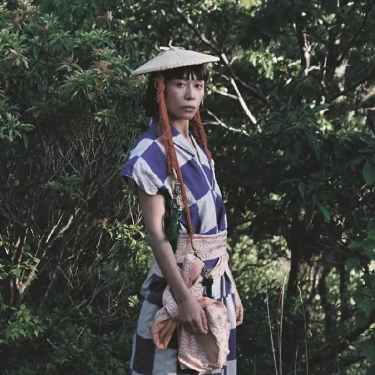 Kina:suttsu stands in front of a bushy backdrop