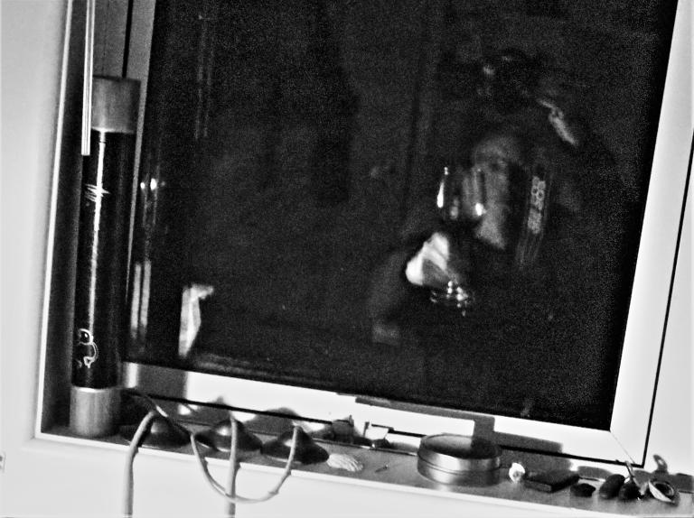 A blurred black and white photo, very grainy. There are chimes on a window ledge. The window is refelcting a figure in motion holding up a glass of red wine in a pose of a toast. The reflection is of the photographer.