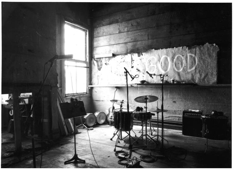 ARC Building Rehearsal Room, black and white photograph of a rehearsal room with one window beaming in bright light into an otherwise very dark room. A drumset, some amps, a music stand and a few mirrors are the only objects in this sparse dingey space.  