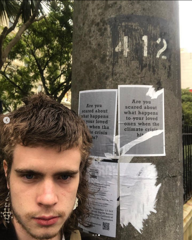 A photo of Baxter in front of a lamp post. The pole has flyers stuck to it, they are slightly torn, and they read "are you ascred about what happens to your loved ones with the climate crisis hits?". Baxter has a mullet and elaborate dangly earrings