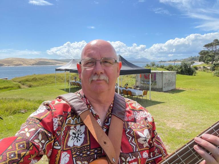 Derek is outside in the sunshine. He is holding a bass. He is bald with grey hair on the edges. He is wearing glasses. The is a gazebo behind him with tables and chairs set up underneath