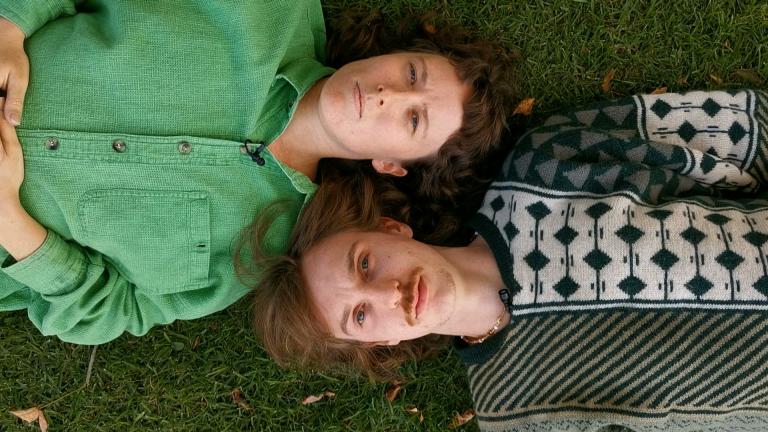 Katie and Oli are lying on the grass lookiing up at the sky. The camera is in the sky looking down at them. Their shoulders are touching and their heads are next to each other. Katie is lying one way and Oli the opposite way.