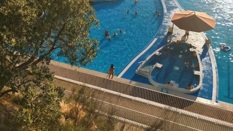 A view down on a pristine blue pool. There are people in the main pool, and also in an inset spa pool. It is clearly a relaxed and beautiful day.