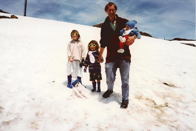 on a mountain of dirty snow stand two children with sunglasses shaped like stars. Their father holds a baby in his arms.