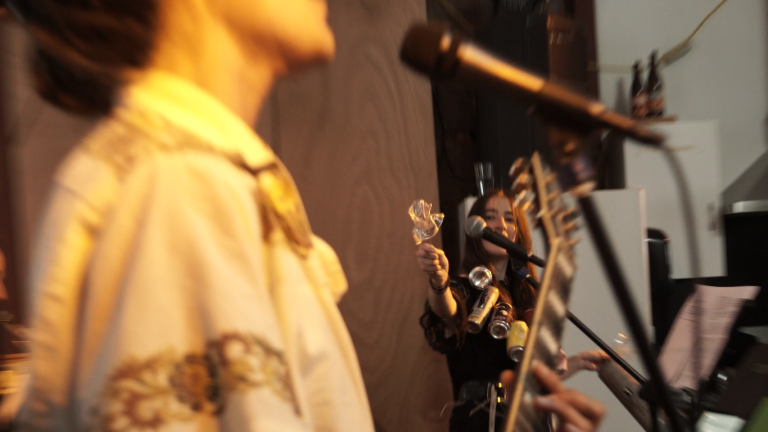 In the foreground is someone playing an electric guitar. In the background someone is playing junk percussion with joy and gusto. The percussion looks to be plastic cups and aluminium cans.