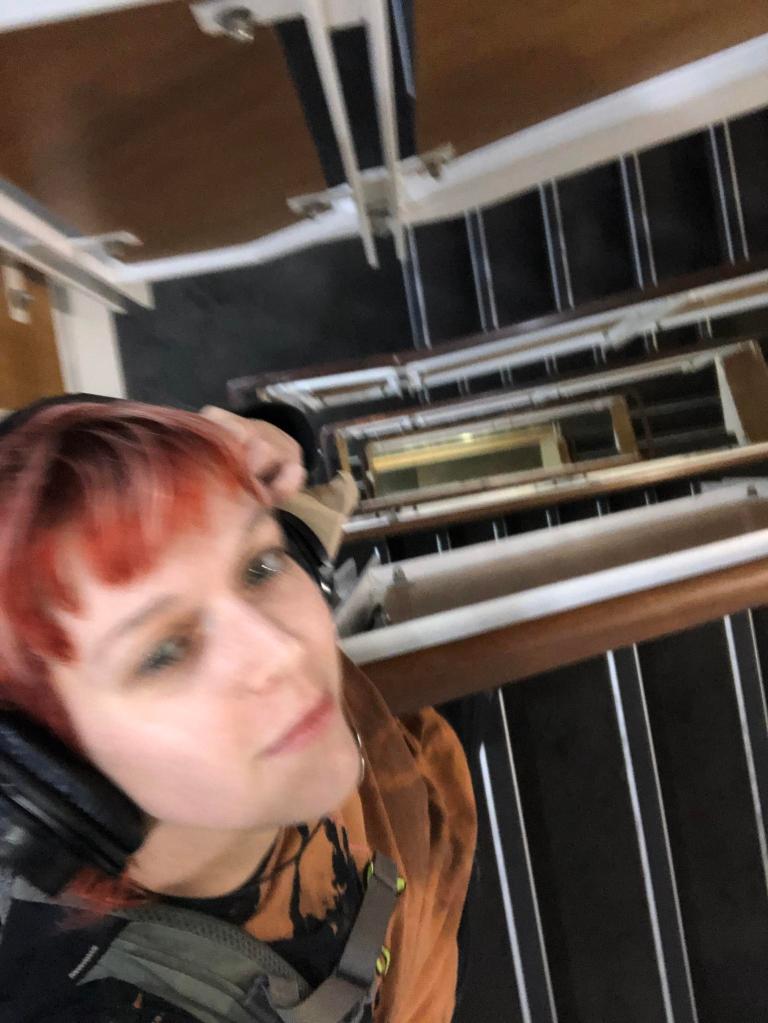 Tash has headphones on and is looking up toward the camera above them. They are at the top of a large stairwell