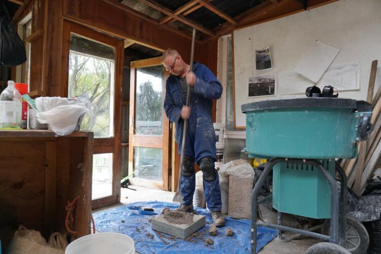 Tim smashing lime stobe with a pole, preparing to make a Roman wall. he is weraing blue overalls, saftey goggles, and is covered in dust.