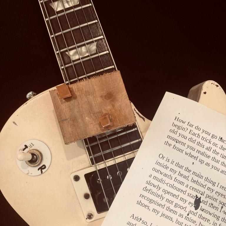 An elctric guitar, Les Paul in style. On top of the strings are the words of Drew's article printed on A4 paper, and a wooden block with rubber squares glued on it.