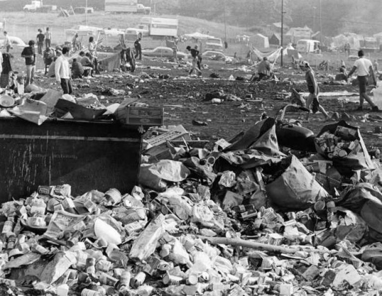 A black and white photo of the afternath of woodstock. There are piles and piles of garbage in what was once a field but is now mud