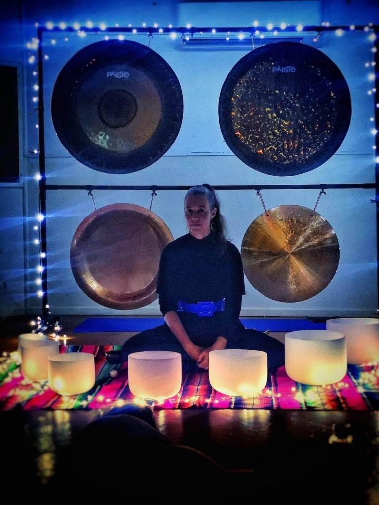 Erika Grant sits cross legged in front of four large gongs that are hanging from a square frame. In front of her there are crystal meditation/prayer bowls that are aglow with warm light