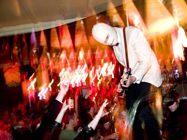 A guitarist wears a white suit, but their head is wrapped in a mask like a Mummy. There is an enthusiastic crowd raising their hands and reaching out in admiration