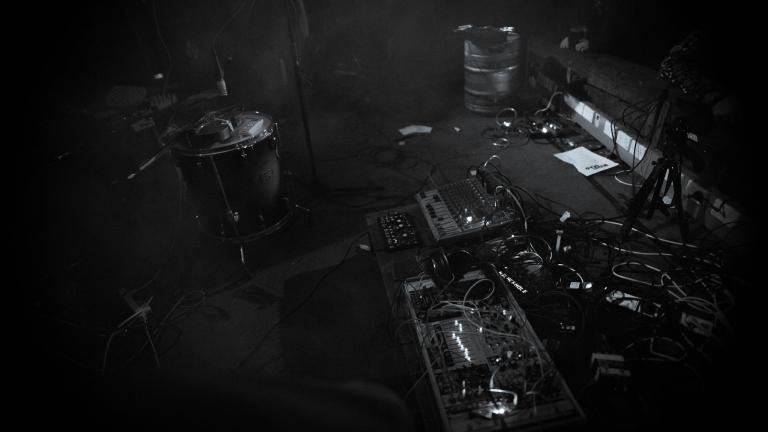 A messy and intriguing set up of cables and boxes and lights. A make shift noise synth. It is in bold black and white. There is a filthy carpet, an empty beer keg, and a dismantled drum kit, in the background.