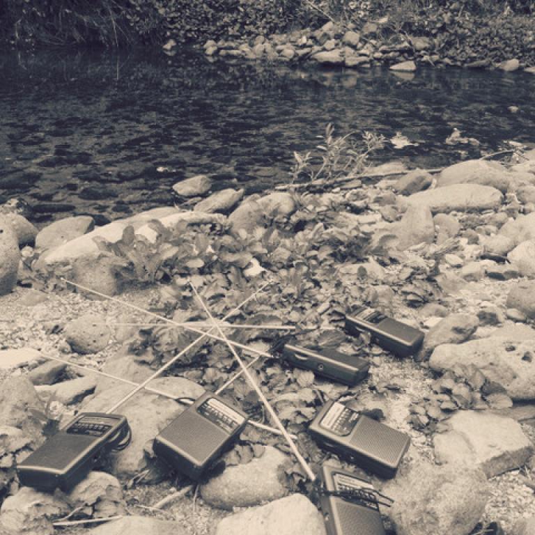 six cheap transistor radios lie around on a river bank. They are scattered amongst the rocks, and their aerials are touching, tangled together. The river flows beside them.