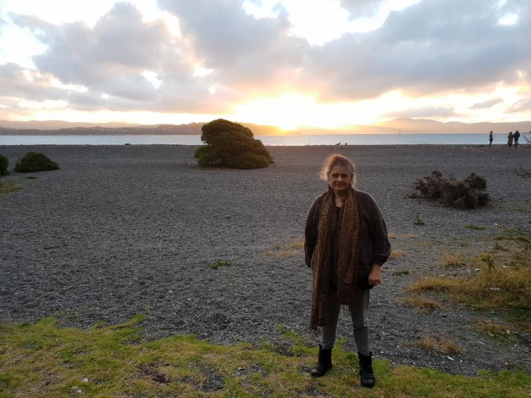 Athena is standing on a stoney beach. The sun is setting behind her and creating a heavenly glow