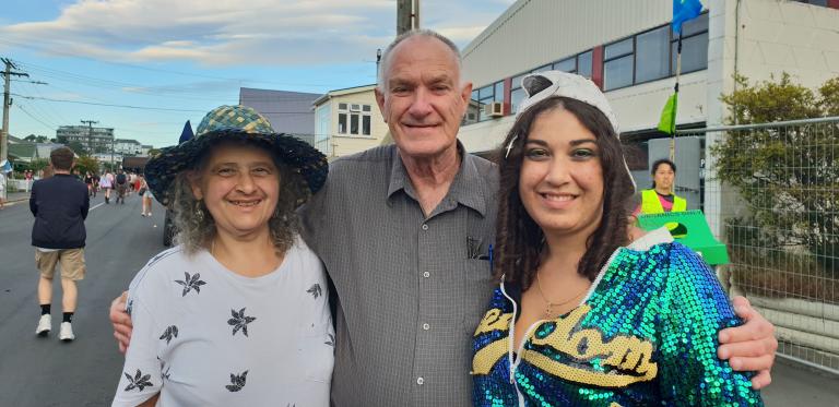 Gerry with his arms around his wife and daughter, Athena and Marika. They are outdoors on a street and the day is clear.