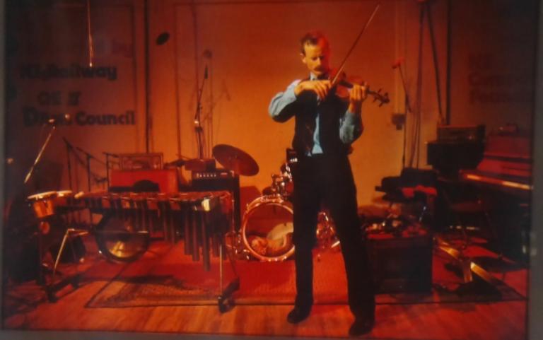 A colour photo of Peter playing. He stands erect and his feet are firmly in place. He is wearing a blue shirt and black waistcoat. The room is red and orange. There is a vibraphone and drum kit behind him, yet he is alone on stage.